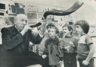 Black and white photo of man holding long spiraled instrument to lips as children look on in cl…
