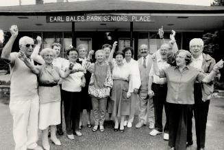 A group of elders stand in front of the Earl Bales Park Senior Place, arms raised in celebratio ...