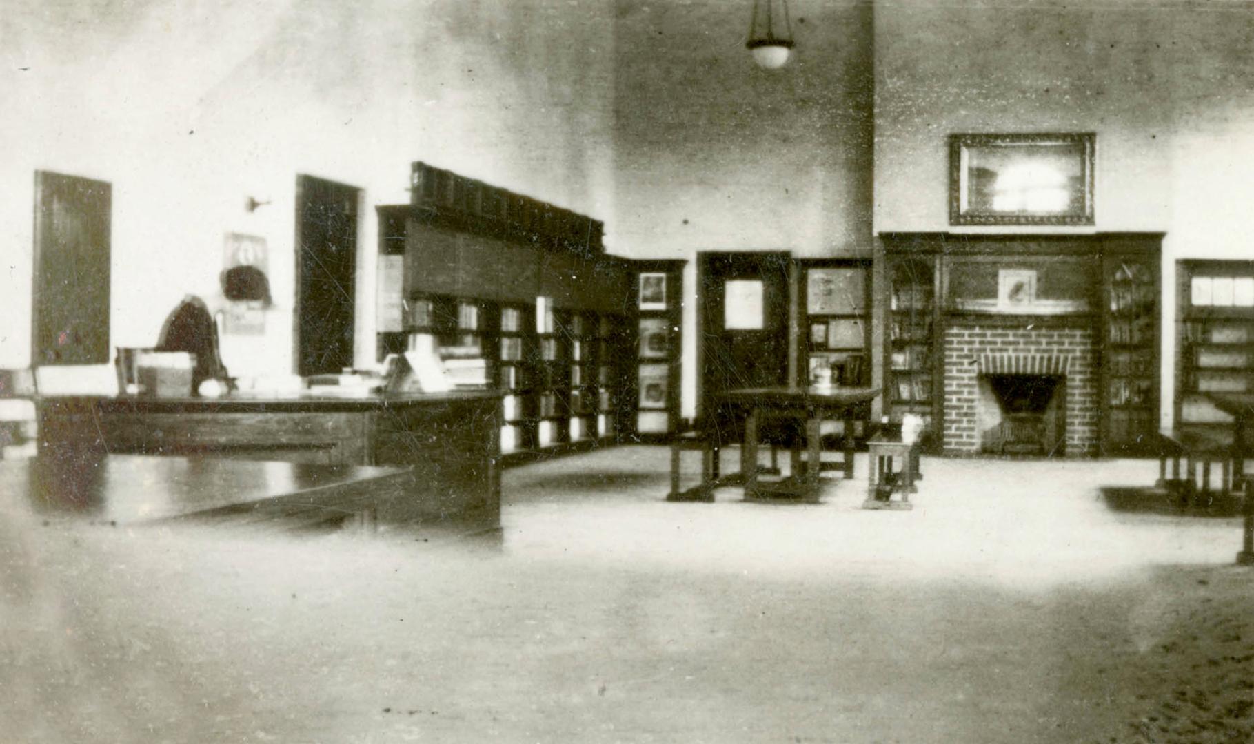 Image shows a library branch interior with some desks, book stacks and a fireplace. There is st…