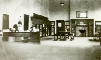 Image shows a library branch interior with some desks, book stacks and a fireplace. There is st…