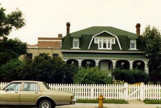 Image shows a two storey residential house with a low fence around it and an automobile parked  ...