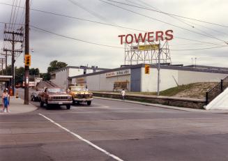 Image shows a street view with some cars on it and a building with a sign that reads "Towers".