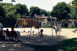 Image shows a few adults and children on the playground with some trees in the background.