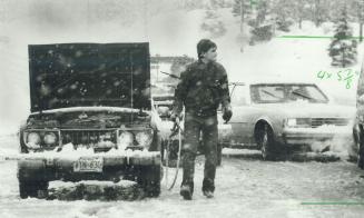 Image shows a motorist, battery booster cables in hand, stands beside his car stalled on Leslie…