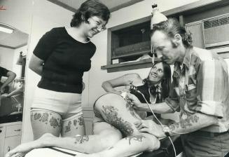 Beachcomber Bill's wife, Brenda, smiles from table as he shows how he applies tattoo