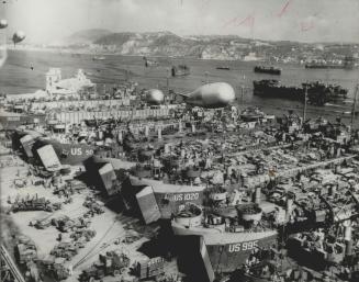 Barrage balloons ride overhead as a line of Allied LST's is loaded with vehicles and other supplies at a southern Italian harbor in preparation for th(...)