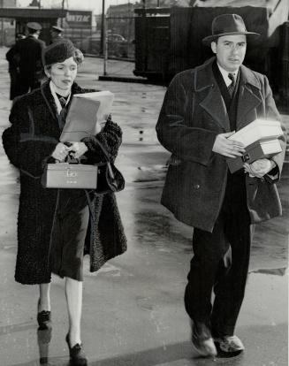A woman in a fur coat, pillbox hat, and a man in a fedora carry documents.