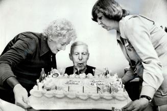 Image shows a gentleman blowing candles on a big birthday cake held by two ladies.