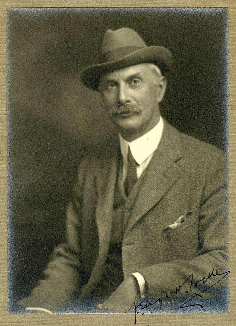 Sepia-toned studio portrait of an adult individual seated and turned slightly to the side, wear…