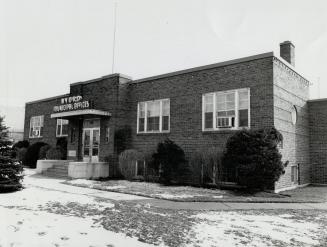 Exterior view of one-storey, flat-roofed brick building with shrubs planted either side of entr…