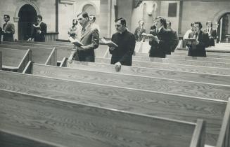 Men dressed in suits (one in religious habit) stand sparsely occupied pews with songbooks in ha ...