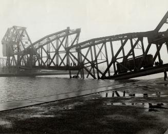 A train trestle is broken in two, collapsed into a river, with a single train car and several p ...
