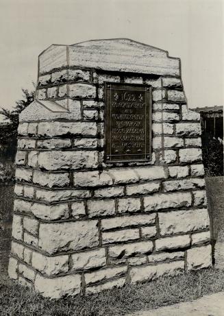 Shows a stone monument with a plaque, standing next to a sidewalk.