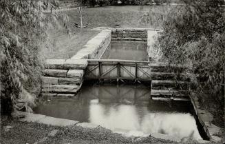 A fragment of a narrow wooden lock, an anchor stands next to it. Bushes grow near the edges in …