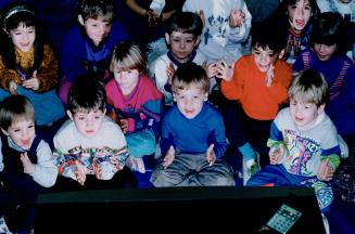 A group of very young children sit cross-legged, clapping their hands and smiling.