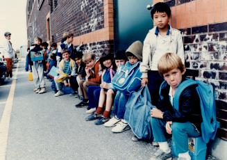 Lunch-pail brigade: Grade 1 students line up, sitting on their lunch boxes, to await a school bus at Agnes Macphail Public School