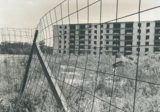 Seen beyond a wire fence supported by wooden beams on unmown grassy land, is a six-storey brick…