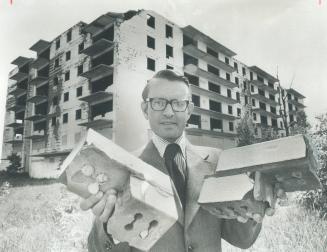 In foreground, man in suit with dark rimmed glasses looks toward camera while holding up buildi…