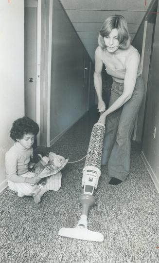 A woman vacuums a hallway while a six-year-old girl watches.