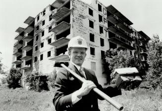Man wearing suit and hardhat smiles as he rests sledgehammer over shoulder and holds up brick;  ...