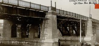 View from below of a large bridge, showing cement support pillars and steel arches. Decorative  ...