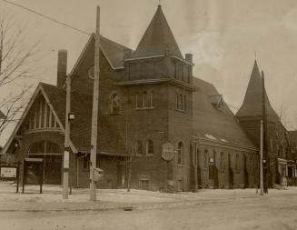 Exterior of brick church with steep gable roof and attached towers front and rear. Front gabled…