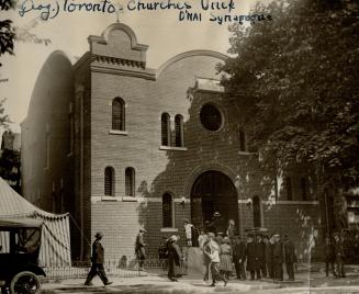 A small group of men, women and children congregate at the entrance steps of a synagogue. 