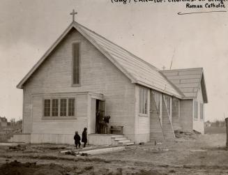 Exterior of unadorned, white clapboard church with small cross affixed to roof and several entr ...