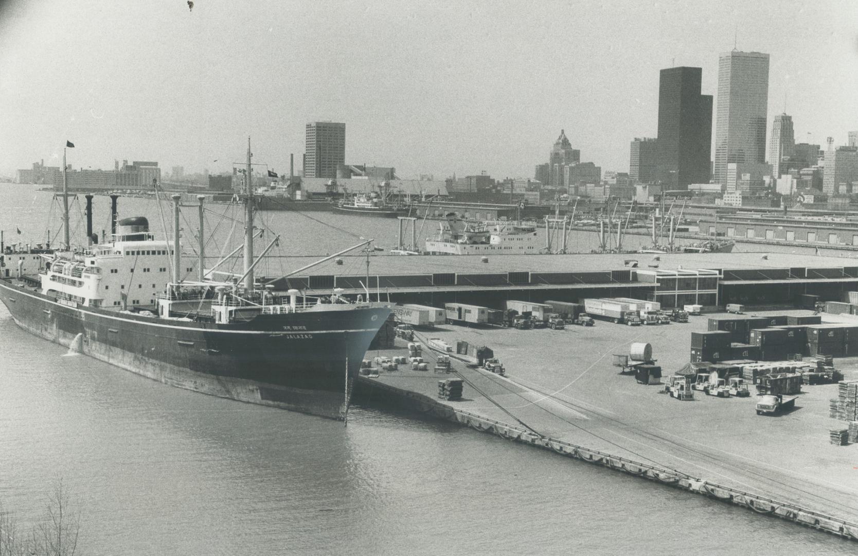 Image shows a big ship docked at the Harbour with some buildings in the background.