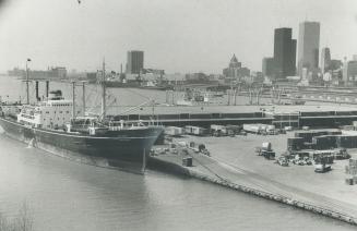 Image shows a big ship docked at the Harbour with some buildings in the background.