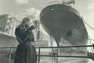 Image shows a young lady standing at the Harbour with a few cargo ships in the background.