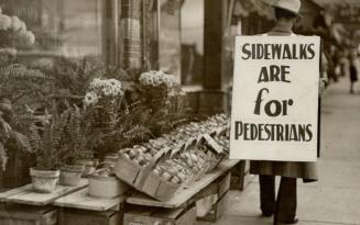Man wearing a protest sign, "Sidewalks are for pedestrians," outside of McGuinness Florist, Danforth Avenue, northeast corner of Woodmount Avenue, Toronto, Ontario
