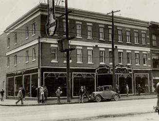 Street view of three-storey brick building at intersection with large shop windows. Sign above  ...