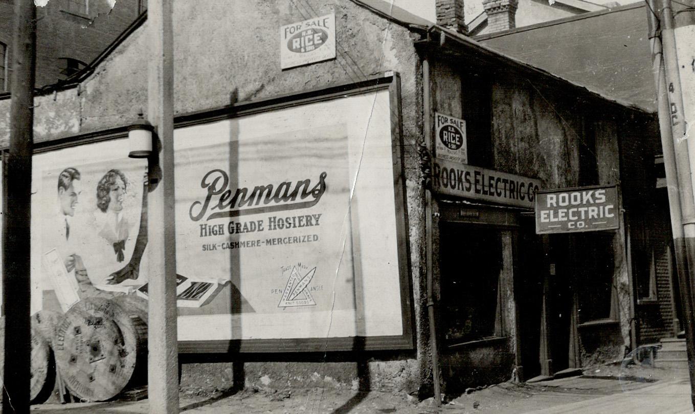Side view of a two-storey building in state of disrepair with large Penman's Hosiery billboard …