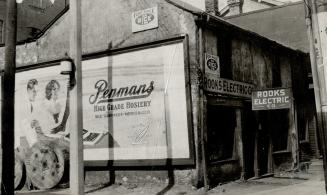 Side view of a two-storey building in state of disrepair with large Penman's Hosiery billboard …
