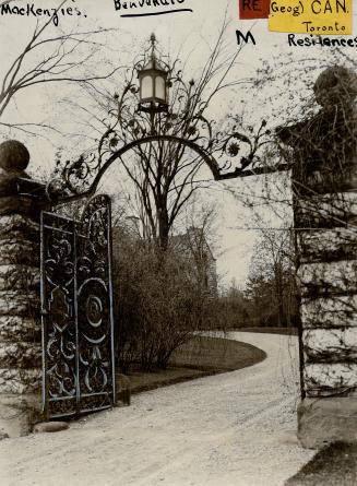 Chiseled stone posts support a decorative wrought iron arched gate at the entrance of a gravel  ...
