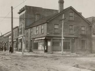 Street view of three-storey brick building with gable roof at corner of intersection. Shop wind ...