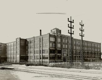 Four-storey brick building; train tracks and two hydro towers seen in foreground.