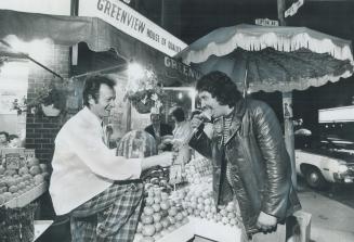 Standing at a fruit and vegetable stand covered by a large fringed umbrella, a man in plaid pan…
