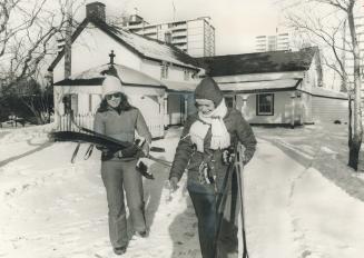 Two women carrying cross-country skis in front of an historic home. High-rise buldings can be s ...