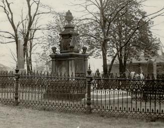 Large stone monument topped with carved urn stands beyond decorative iron fence.