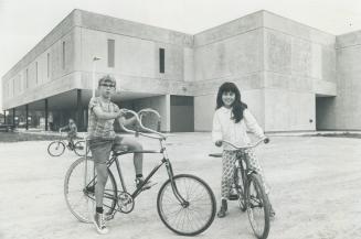 Boy and girl on bikes stand in front of concrete, two-storey building with narrow, slit windows ...