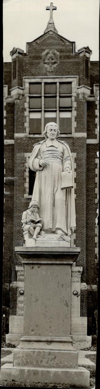 Carved, white stone statue of a man in gown and robe, holding paper in hand; left, a statue of  ...