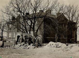 Men with shovels stand near mounds of dirt; beyond is a two-storey brick building.