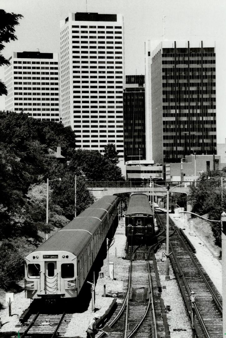 Image shows a few subway trains on tracks outdoors and and a number of high-rise buildings in t ...