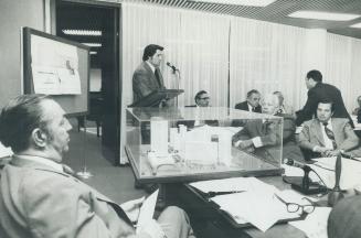 Image shows a person speaking into the microphone and others listening at the desks covered wit ...
