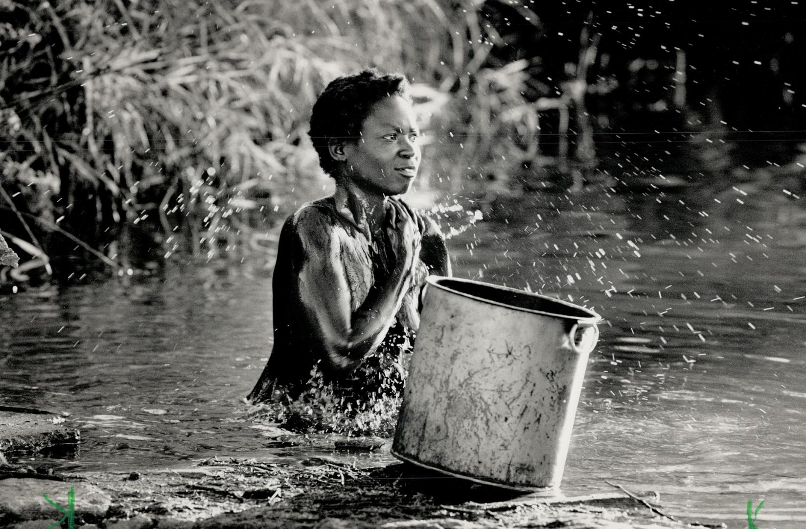 Kneeling in a nearby stream, a village girl showers herself with an aluminium pot