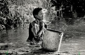 Kneeling in a nearby stream, a village girl showers herself with an aluminium pot