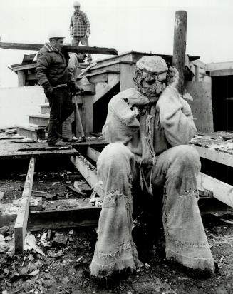 Actor in costume sits on the edge of a stage that workmen, in the background, are dismantling.