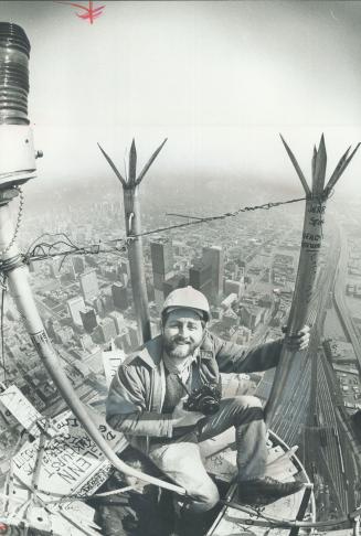 Image shows a photographer at the top of the CN Tower with an aerial view of Toronto in the bac ...